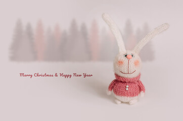 Handmade knitted bunnies stand against a white background. Behind is a forest of fir trees. Postcard for congratulations on the New Year and Christmas holiday.