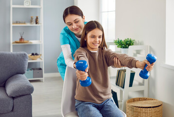 Child girl doing exercise using dumbbells with support from nurse.Young smiling caregiver...