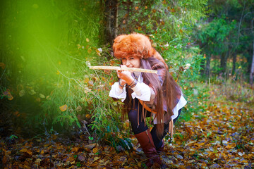 Girl in a leather jacket, a big red fox fur hat and with a crossbow in the forest in autumn. A...