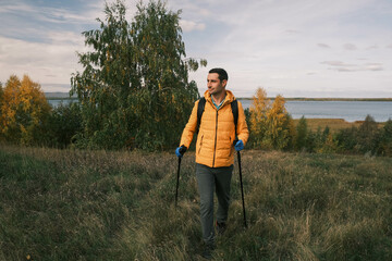 Fototapeta premium man with a backpack and gloves is walking with trekking poles on an autumn field against the background of the lake.An active lifestyle.Walking with trekking poles