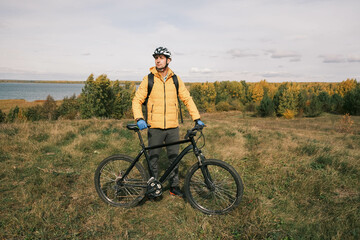 Obraz premium man in a protective helmet and a backpack stands next to a mountain bike in autumn against the background of a forest and a lake.An active lifestyle.Cycling