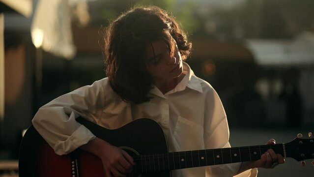 Beautiful girl plays an acoustic guitar while sitting on a chair in the middle of a crowded street Positive guitarist moving head during song Musician performing musical composition on guitar outdoors