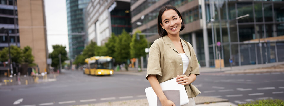 Beautiful asian girl smiles as commutes to work, stands on street with laptop and notebook