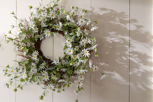 Wreath with green leaves and white and yellow daisy flowers hanging on white wood panel wall with shadow from sun shining through window