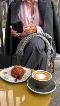 Woman sitting in outdoor cafe, browsing smartphone or sending message, picks up cup of cappuccino coffee in her hands and croissant on table comfortably wrapped in gray plaid scarf.