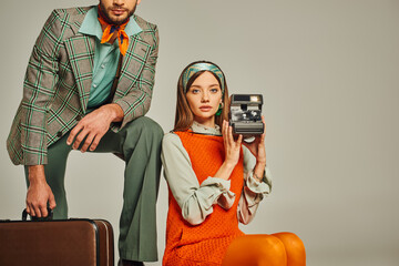 woman in orange dress holding vintage camera near man with suitcase on grey, retro fashion