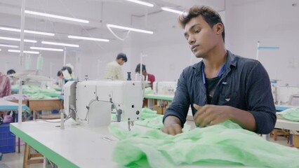 A skilled Indian young man or male artisan or tailor is stitching a dress on a sewing machine along with other workers in a busy cotton or textile workshop or garment industry or factory