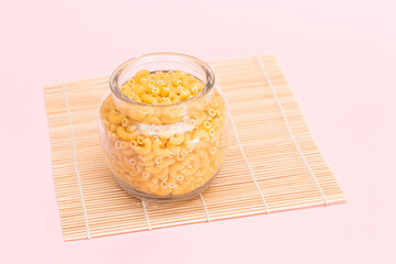 Uncooked Chifferi Rigati Pasta in Glass Jar on Bamboo Mat on Pink Background. Fat and Unhealthy Food. Scattered Classic Dry Macaroni. Italian Culture and Cuisine. Raw Pasta