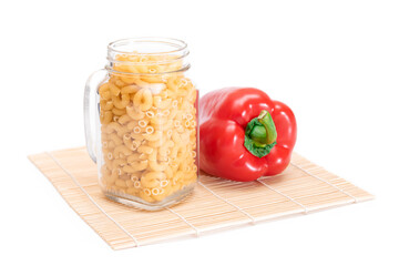 Uncooked Chifferi Rigati Pasta in Glass Jar and Red Bell Pepper on Bamboo Mat Isolated on White Background. Fat and Unhealthy Food. Classic Dry Macaroni. Italian Culture and Cuisine. Raw Pasta