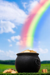 A black cast iron pot full of gold coins sitting in the grass at the end of a rainbow. Some gold coins are stacked on the grass next to the pot. 