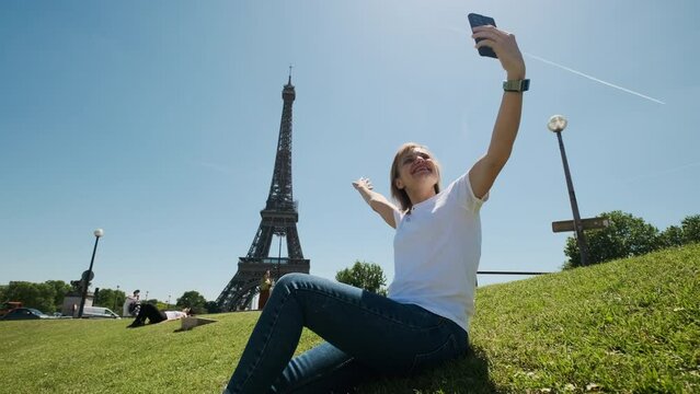Woman is taking selfie using smartphone sitting near Eiffel tower in Paris in daytime, Wide footage