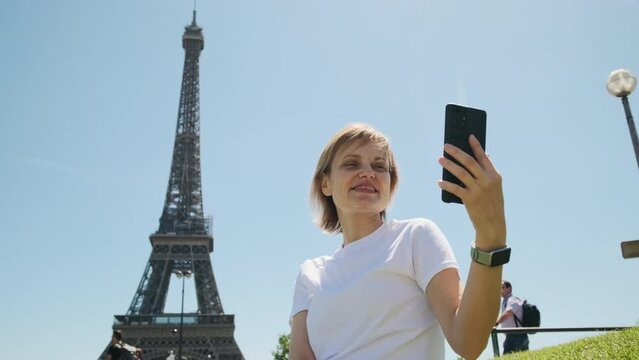 Woman is taking selfie using smartphone sitting near Eiffel tower in Paris in daytime, Close up view