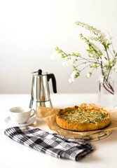 There is a quiche pie on parchment paper, next to it there is a coffee pot and a white cup and saucer. There is a vase of white flowers on the table and a black and white checkered napkin