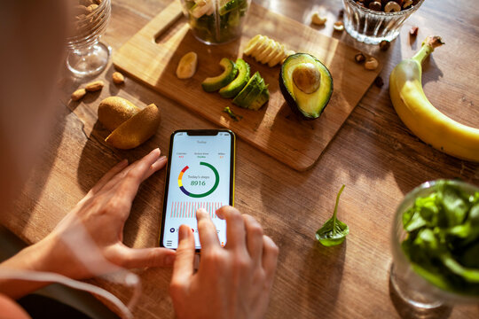 Young woman checking the health app on her smartphone while making a healthy smoothie in the kitchen