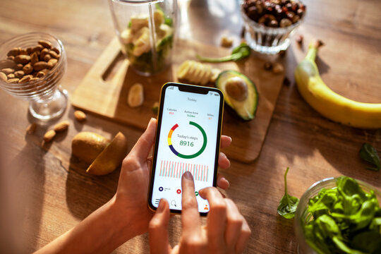 Young woman checking the health app on her smartphone while making a healthy smoothie in the kitchen