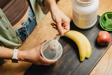 Woman in jeans and shirt with a measuring spoon in her hand puts portion of whey protein powder...