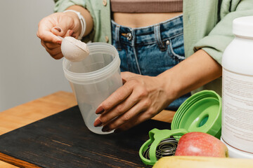 Woman in jeans and shirt with a measuring spoon in her hand puts portion of whey protein powder into a shaker on wooden table with white capsules, bananas and apple. Process of making protein drink