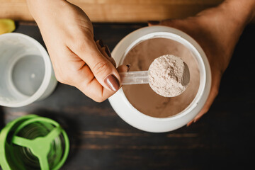 Female hand holds measuring spoon with portion of whey protein powder above plastic jar on wooden...