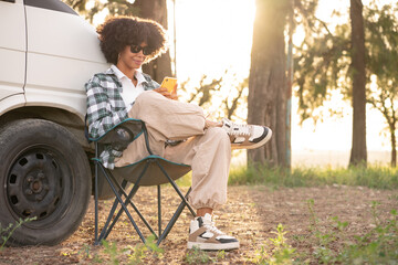 Young African black woman in the nature using her smartphone during a road trip in her camper van...