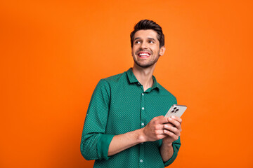 Photo of cool dreamy man dressed green shirt texting modern gadget looking empty space isolated orange color background