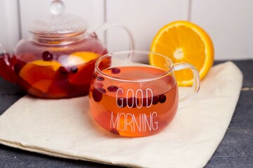 Red berry tea in a transparent mug on a white linen napkin, next to a transparent teapot with tea and an orange.