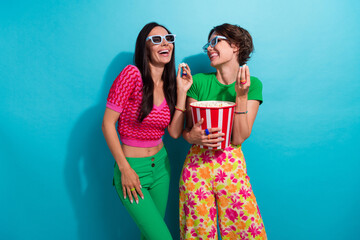 Photo of optimistic two young girlfriends laughing enjoy watching 3d cartoons eating biting popcorn isolated on blue color background - obrazy, fototapety, plakaty