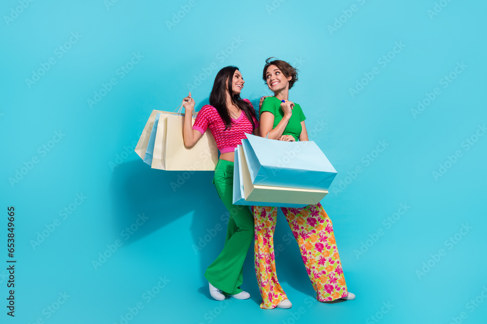 Wall mural Full size photo of cheerful beautiful two girlfriends hold packages shopping together make happiness isolated on blue color background