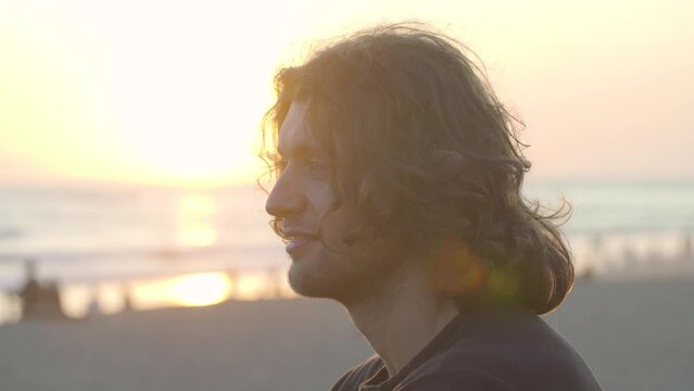 Close-up of young adult handsome man with long curly hair and stubble looks forward at sunset beach 4K
