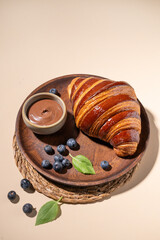 Freshly baked croissant with chocolate on a wooden plate and blueberries on a yellow background. The concept of a delicious homemade breakfast.