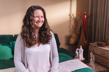 Beautiful woman with wavy hair, smiling, sitting on bed wearing pajamas.