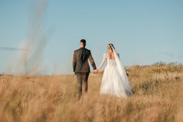 shot of a beautiful bride in a white dress and the groom holding her hand in the middle of a sunny day in the field, wedding photography, newlyweds walking together, love,common future