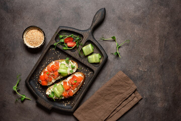 Open sandwich with salmon, cucumber and microgreens on a wooden cutting board, dark rustic background. Top view, flat lay, copy space.