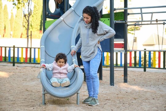 Dominican mother playing with her toddler daughter at the slide in a park. Latin family.