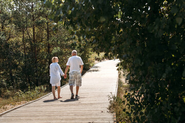 Fototapeta premium Happy elderly couple walking on a wooden deck on a forest path in the sand dunes