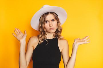 Discontent disgruntled young blonde woman spreading hands scream shout mouth wide open isolated on yellow background studio portrait. Yelling, quarrel concept. Girl wear white hat, black top. - obrazy, fototapety, plakaty