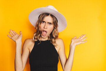 Discontent disgruntled young blonde woman spreading hands scream shout mouth wide open isolated on yellow background studio portrait. Yelling, quarrel concept. Girl wear white hat, black top. - obrazy, fototapety, plakaty