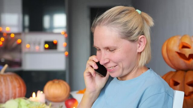 Сlose up of laughing pretty young woman talking on cell phone while sitting in modern kitchen against background pumpkins Halloween festival decorations lanterns Jack. Invitation autumn carnival.