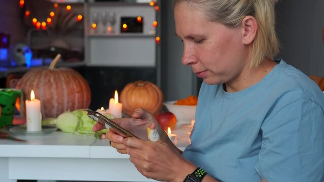 Young happy woman using mobile phone against background of holiday decorations during Halloween, surrounded by glowing garlands of burnt candles and traditional large pumpkins, in kitchen at home.