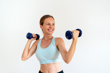 Young female brunette female wearing sportswear holding two dumbbells in her hands in front of white background.