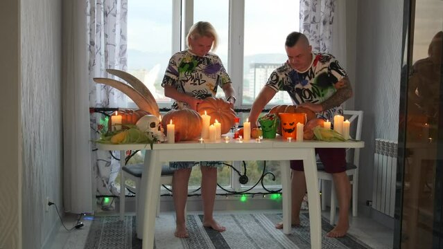 Guy and a girl in same T-shirts in the kitchen, decorated with glowing garlands, enthusiastically carve Jack-o'-lantern pumpkins preparing for the fall holiday in the Halloween festival, background.