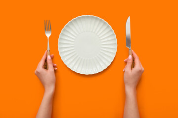 Female hands with cutlery and empty plate on orange background