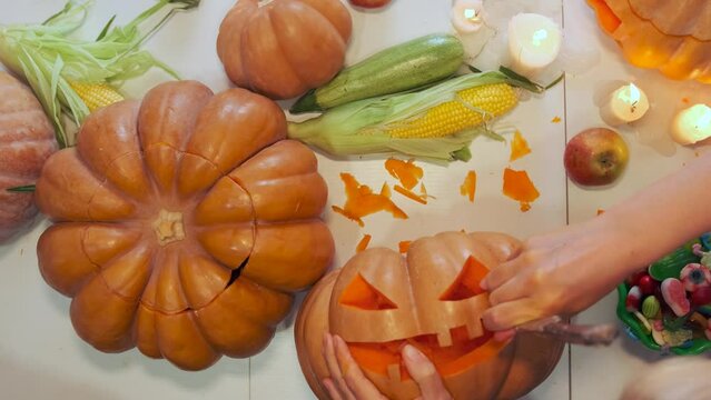Top view of festive background girl's hands carving with knife creepy jack o' lantern face on table with peeled traditional pumpkins corn apples and candies. Happy holiday during Halloween. Close up.