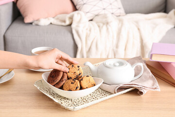 Woman holding cookie and teapot with cup of tea on wooden table in living room