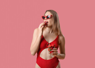 Young woman in swimsuit eating strawberries on pink background