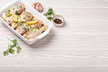Raw uncooked chicken legs with seasonings, herbs, lemon in white ceramic casserole top view on light wooden rustic background. Preparing healthy meal with marinated chicken drumsticks. Space for text.