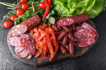 fresh herbs and vegetables, dry-cured sausage, dried pieces of Carpaccio meat, small salami sticks, vintage wooden cutting board, on a black isolated background