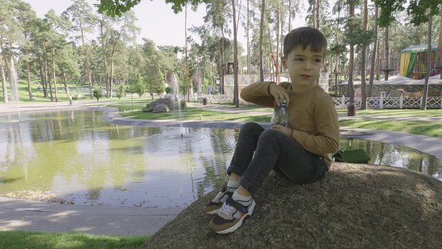 A boy drinks water from a bottle while sitting on a stone in the park.