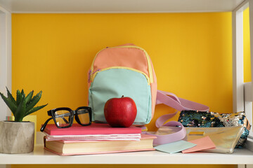 School backpack, stationery, eyeglasses and apple on shelf unit near color wall