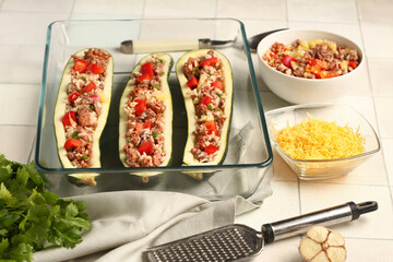 Baking dish with meat stuffed zucchini boats and ingredients on white tile background