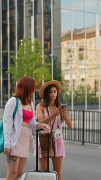 A pair of animated young ladies, each with a suitcase, is walking the city streets and employing their smartphones to find the needed destination.